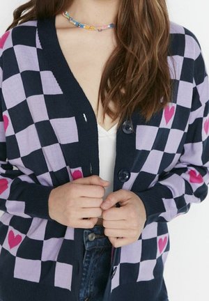 Cardigan avec un motif damier bleu marine et lavande, avec des accents en forme de cœur rose, fermeture à boutons et poignets côtelés. Matière en tricot douce.