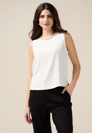 Haut sans manches blanc avec une texture lisse et un col rond orné d'un accent en chaîne dorée ; porté avec un pantalon noir fuselé.