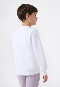 Vit sweatshirt med långa ärmar, rund halsringning och ribbade muddarna och nederkant. Buren över ljuslila byxor, sedd bakifrån.