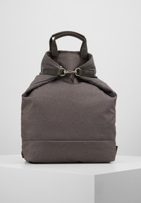 Mochila de tela gris con forma redondeada, que cuenta con un asa superior, correas ajustables y herrajes metálicos para el cierre.