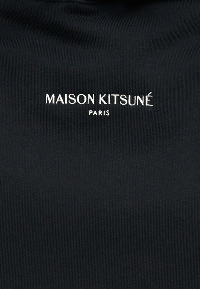 Μαύρο ύφασμα με λευκά κεντημένα γράμματα "MAISON KITSUNÉ" πάνω από μικρότερα γράμματα "PARIS", στο κέντρο του υλικού.