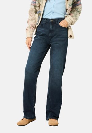 Person trägt dunkelblaue Jeans mit geradem Bein, ein blaues Hemd mit Knöpfen, einen mehrfarbigen gestrickten Cardigan und hellbraune Schuhe.