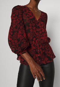Blusa nera con un motivo floreale rosso, scollatura a V, maniche a palloncino e chiusura con bottoni. Tessuto morbido con una texture leggermente trasparente.