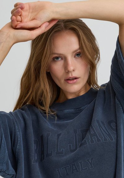 Jeune femme aux longs cheveux châtain clair portant un sweat-shirt ample de couleur foncée avec le texte "BILL JEANS ITALY", les bras levés au-dessus de la tête.