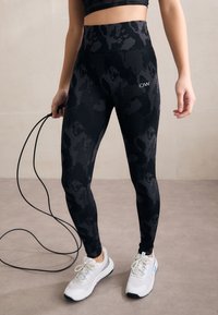 Zwarte hoge taille legging met een camouflagepatroon, gemaakt van rekbare stof. Gecombineerd met witte sportschoenen en een springtouw.