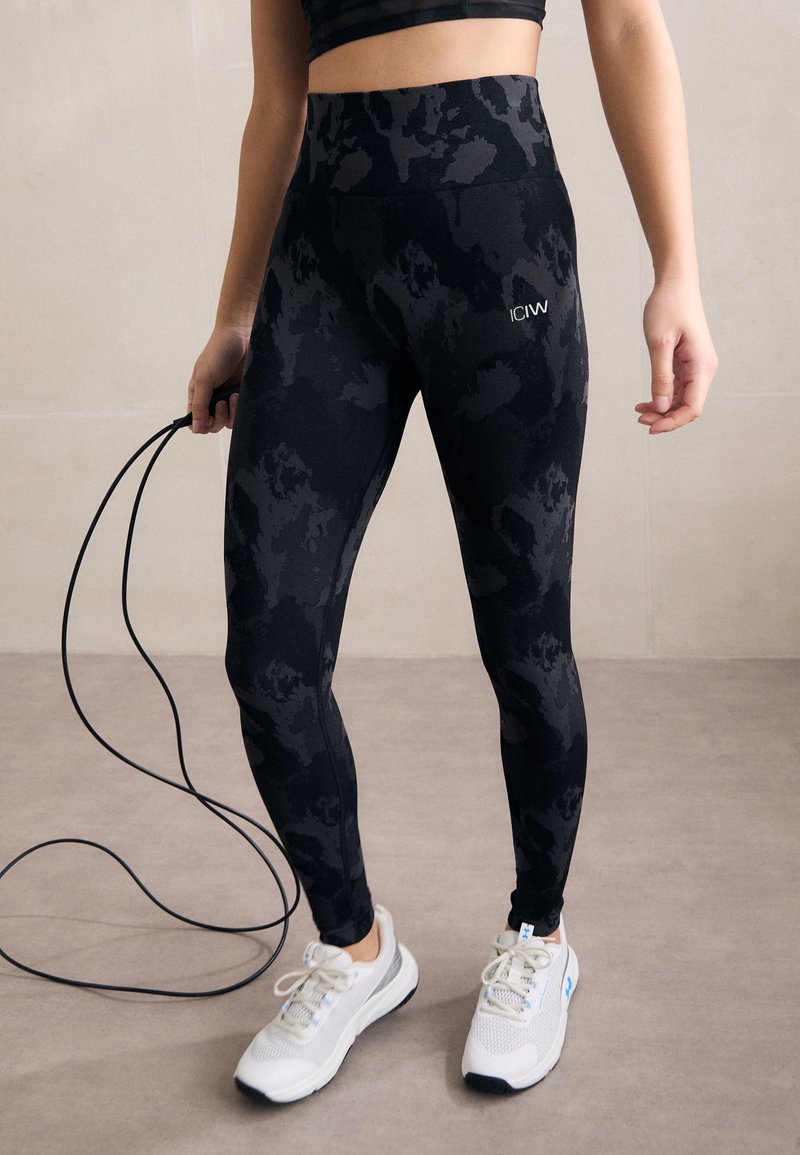 Zwarte hoge taille legging met een camouflagepatroon, gemaakt van rekbare stof. Gecombineerd met witte sportschoenen en een springtouw.