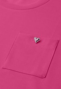 Chemise rose vif avec une poche poitrine assortie et une petite épingle argentée brillante en forme de cœur attachée près du bord supérieur de la poche.