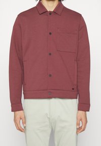 Maroon jacka gjord av mjukt tyg, med knäppning framtill, krage, ett bröstfack och två sidofickor. Slät textur och avslappnad design.