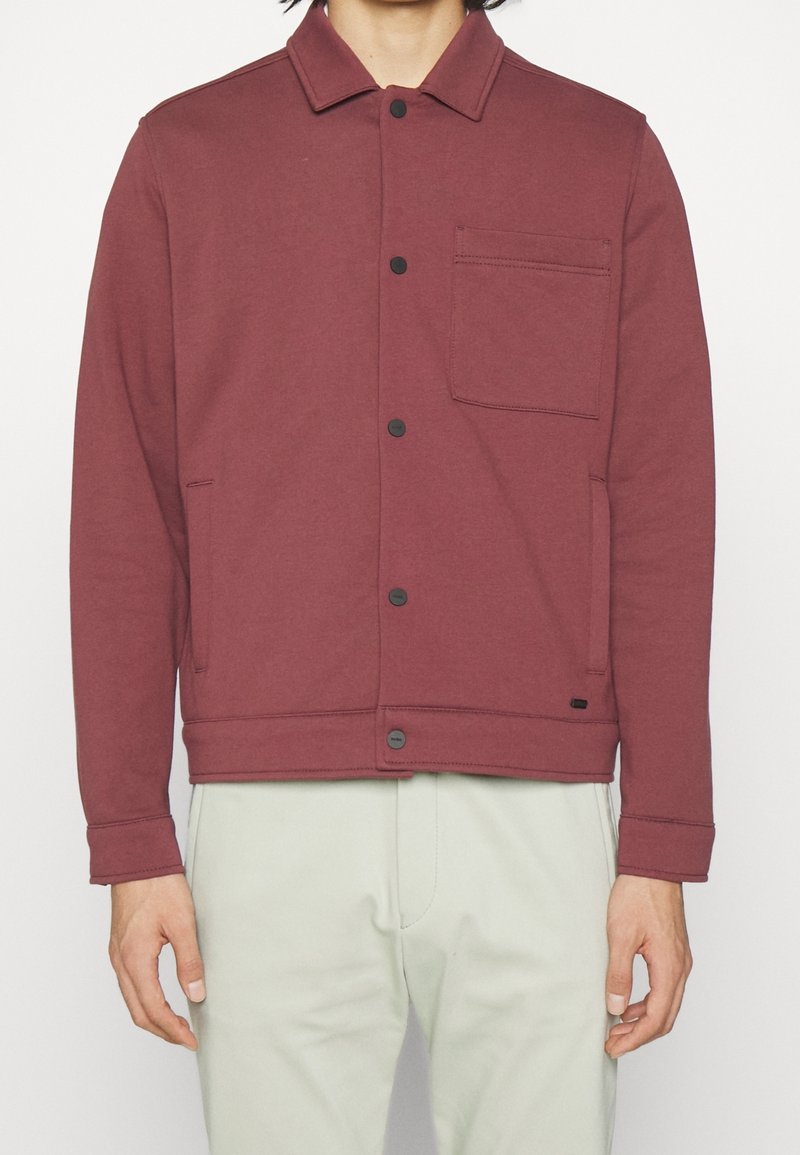 Maroon jacka gjord av mjukt tyg, med knäppning framtill, krage, ett bröstfack och två sidofickor. Slät textur och avslappnad design.