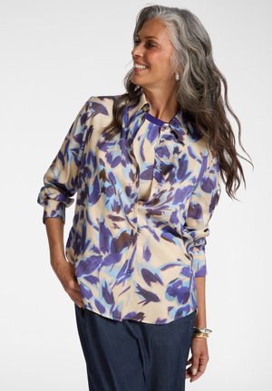 Lächelnde Frau mit langem grauem Haar trägt eine lockere, blaue und beige Blumenbluse, dunkle Hosen und silberne Armbänder, steht mit der Hand in der Tasche.