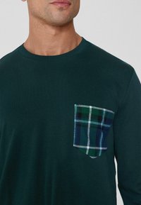 Zielony t-shirt z długim rękawem i okrągłym dekoltem, z kieszenią w kratę w niebiesko-zielono-białym wzorze. Miękka bawełniana tkanina.