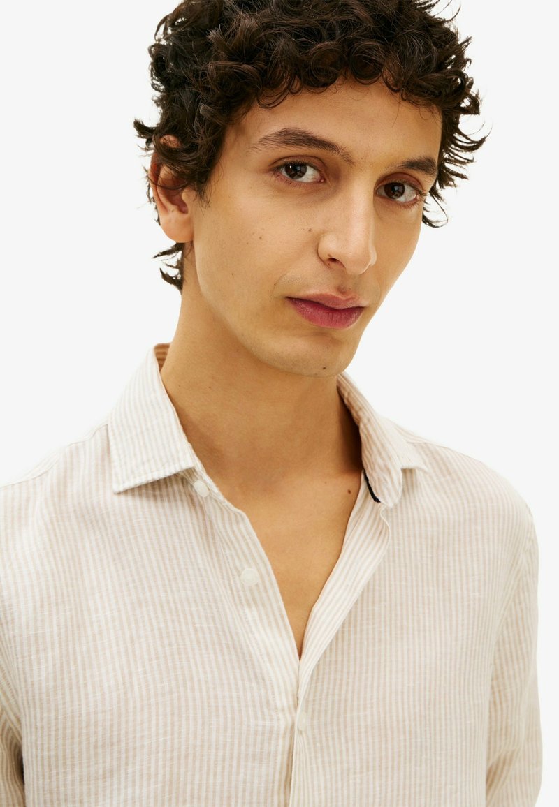 Jeune homme aux cheveux bouclés foncés portant une chemise beige rayée à boutons, regardant légèrement sur le côté sur fond blanc.