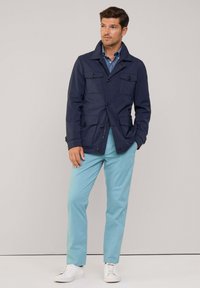 Giacca leggera blu navy con multiple tasche anteriori, dettagli con bottoni e colletto, abbinata a pantaloni azzurri chiari e sneakers bianche.