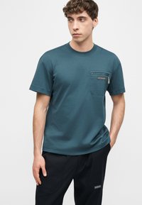 Νεαρός άνδρας φορώντας πετρόλ κοντομάνικη μπλούζα με φερμουάρ στην τσέπη στο στήθος και μαύρο παντελόνι, στέκεται με το ένα χέρι στην τσέπη, κοιτάζοντας πλάγια.