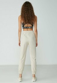 Beige Jogginghosen mit elastischen Bündchen, aus weichem Stoff, kombiniert mit einem schwarzen Bralette mit einem Etikettendesign. Rückansicht.