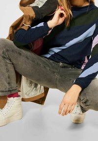 Osoba ubrana w sweter w wielokolorowe paski, szare dżinsy, białe sneakersy na wysokiej podeszwie i brązową kurtkę, klęczy z szeroko rozstawionymi nogami na szarym podłodze.