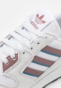 Valkoiset Adidas-tennarit valkoisilla nauhoilla, vaaleanpunaiset nahka- ja samettikoristeet, sekä siniset ja vaaleanpunaiset raidat sivuilla.