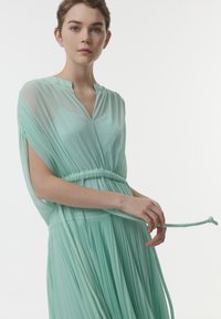 Vestido fluido de color verde menta con tejido transparente, diseño plisado, escote en V y cintura ceñida. Presenta un detalle de ajuste con lazo.