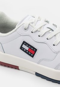 Białe skórzane sneakersy z wygodnym wnętrzem, teksturowanymi podeszwami i czarną naszywką z logo z czerwonymi i niebieskimi akcentami. Buty mają białe sznurówki.
