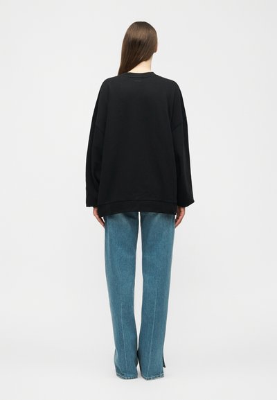 Ember, aki háttal áll, laza fekete hosszú ujjú felsőt és egyenes szabású kék farmert visel, egyszínű fehér háttér előtt.