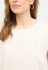 Hellbeige Kurzarmtop mit Rundhalsausschnitt, versehen mit einem dezenten goldenen Pferdelogo auf der Brust. Glatte, weiche Stofftextur.