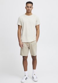 T-shirt beige en coton à manches courtes avec logo, associé à un short beige à cordon de serrage, porté avec des chaussettes blanches et des baskets avec accents noirs.
