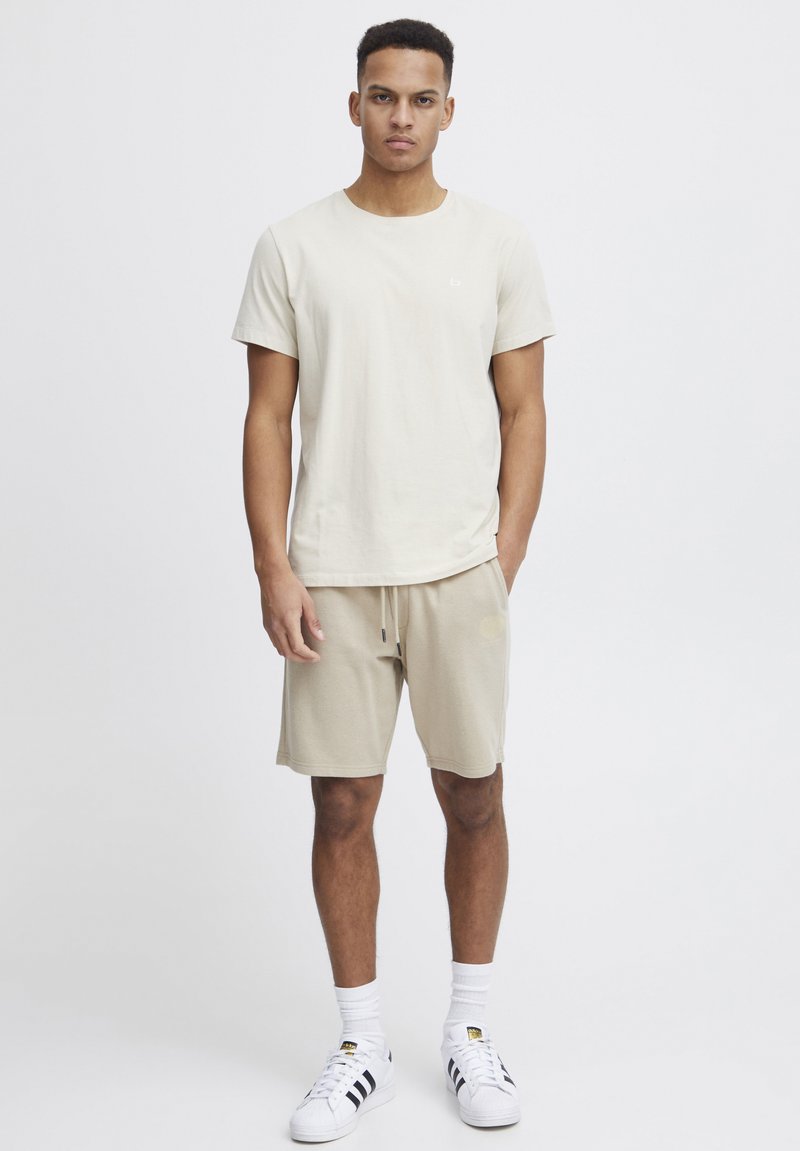 T-shirt beige en coton à manches courtes avec logo, associé à un short beige à cordon de serrage, porté avec des chaussettes blanches et des baskets avec accents noirs.