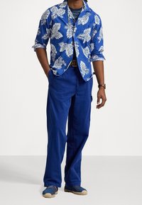 Niebieska koszula z krótkim rękawem w białe liście, w połączeniu z niebieskimi spodniami typu cargo i niebieskimi espadrylami. Koszula ma kołnierzyk na guziki i podwinięte rękawy.