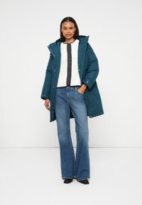 Parka acolchoada teal com capuz, combinada com um suéter branco texturizado e jeans azuis boca de sino. Modelo em pé num fundo neutro.