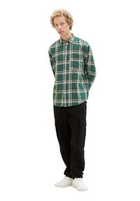 Grünes karriert Button-up-Hemd mit weißen und gelben Akzenten, getragen mit schwarzen Cargo-Hosen und weißen Turnschuhen. Modell steht mit den Händen hinter dem Rücken.