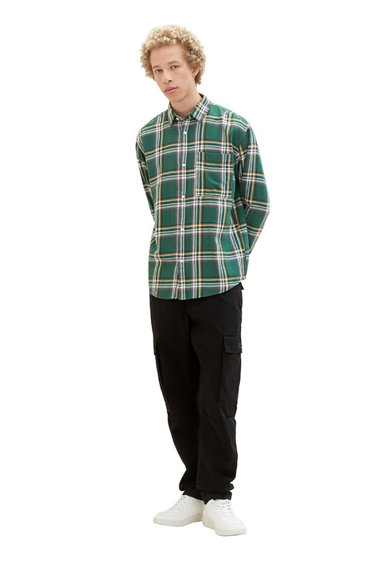 Grünes karriert Button-up-Hemd mit weißen und gelben Akzenten, getragen mit schwarzen Cargo-Hosen und weißen Turnschuhen. Modell steht mit den Händen hinter dem Rücken.