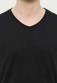 Άνδρας που φοράει απλό μαύρο T-shirt με V-neck, εμφανιζόμενο από τον λαιμό έως το άνω μέρος του στήθους, με ουδέτερο φόντο.