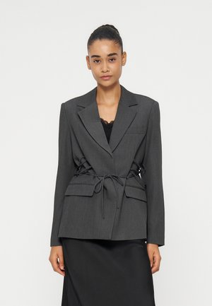 Mujer con cabello oscuro recogido en un moño, lleva un blazer entallado de color gris oscuro atado a la cintura sobre una camiseta y falda negras, sobre un fondo liso.