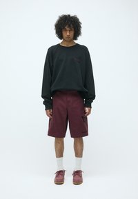 Sudadera negra con logo en burdeos, pantalones cortos burdeos con bolsillos laterales, calcetines blancos y zapatillas burdeos con suelas gruesas beige.