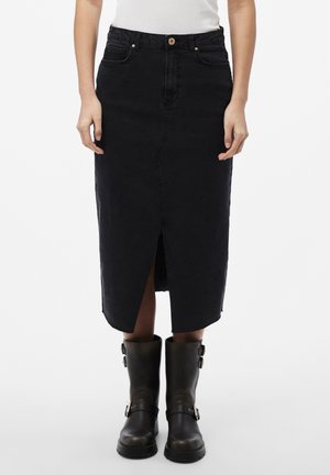 Woman wearing a black denim midi skirt with front slit and dark brown buckle boots, standing against a white background.