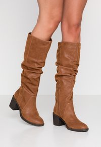 Brown, knee-high slouch boots made of soft leather-like material, featuring a chunky heel and a side zipper for ease of wear.