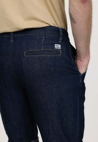 Jeans en denim bleu foncé avec une coupe ajustée, présentant des coutures jaunes, une poche arrière avec une étiquette de marque, et une légère texture.