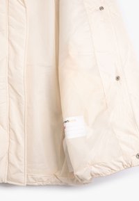 Hellbeige Pufferjacke innen; verfügt über ein gesteppte Design, weiche Textur, Druckknöpfe und ein Etikett mit Platz zum Schreiben von Namen.