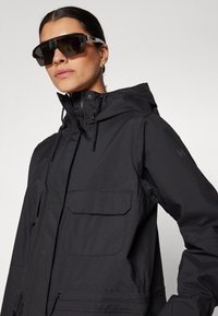 Zwarte waterdichte jas met een capuchon, voorzien van twee voorzakken en koorddetails, gedragen met oversized zwarte zonnebril.