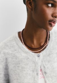 Geschichtete Halsketten mit dünnen Strängen in Pink, Burgunder und Grün, getragen mit einer hellgrauen, flauschigen Strickjacke und einem weißen Hemd.