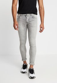 Hombre que lleva puestos unos jeans ajustados en gris claro con puños, una camiseta negra y zapatillas Nike blancas, de pie contra un fondo liso.