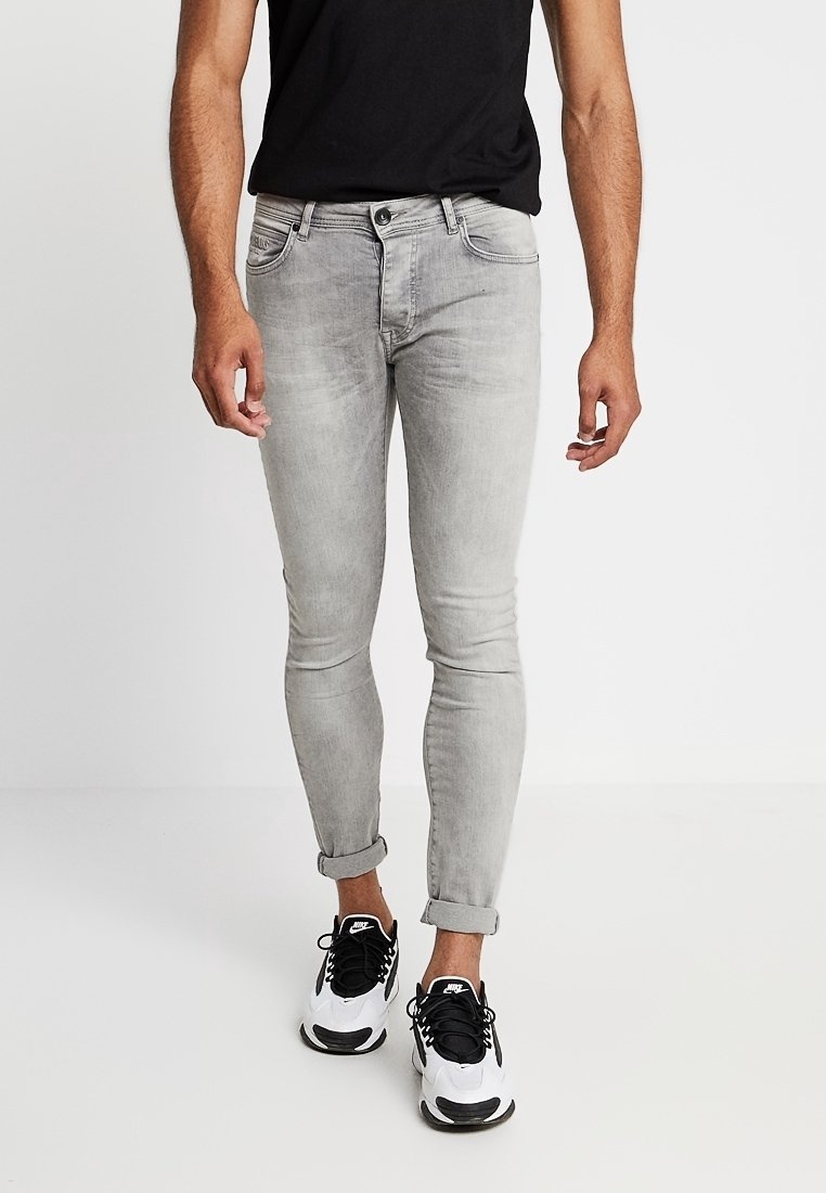 Hombre que lleva puestos unos jeans ajustados en gris claro con puños, una camiseta negra y zapatillas Nike blancas, de pie contra un fondo liso.