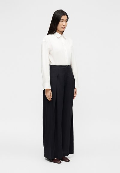 Γυναίκα με μακριά μαύρα μαλλιά στέκεται, φορώντας λευκό πουκάμισο με κουμπιά, φαρδιά μαύρη παντελόνα και σκούρα καφέ παπούτσια, μπροστά σε απλό φόντο.