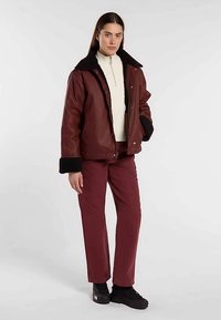 Brun læderjakke med sort pelskant, kombineret med en hvid top med høj hals og burgunderbukser. Modellen har sorte sneakers på.