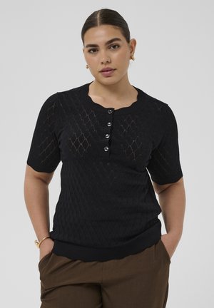 Kvinde med mørkt hår iført en sort kortærmet tekstureret knapbluse og brune bukser, stående med hænderne i lommerne på grå baggrund.