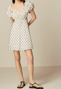 Vestido de manga corta en crema con patrones negros, cintura ajustada y falda acampanada; combinado con sandalias planas negras. Textura de tela suave.