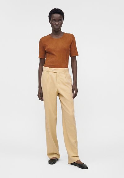 Γυναίκα όρθια με μπλούζα κοντομάνικη ριμπ σε σκουριά απόχρωση, μπεζ πλισέ παντελόνα με φαρδύ πόδι και μαύρα επίπεδα παπούτσια.