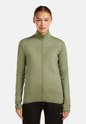 Femme portant une veste olive à manches longues avec fermeture éclair et passages pour les pouces, debout contre un fond blanc uni.