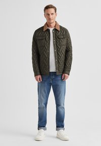 Steppjacke in Olivgrün mit braunem Cordkragen, zwei Brusttaschen, Druckknöpfen, kombiniert mit blauen Jeans und weißen Turnschuhen.