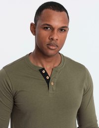 Man med kort hår som bär en olivgrön långärmad henley-shirt med tre beige knappar mot en enkel ljus bakgrund.